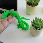Dragão verde impresso em 3D sendo segurado na mão, com plantas suculentas ao fundo