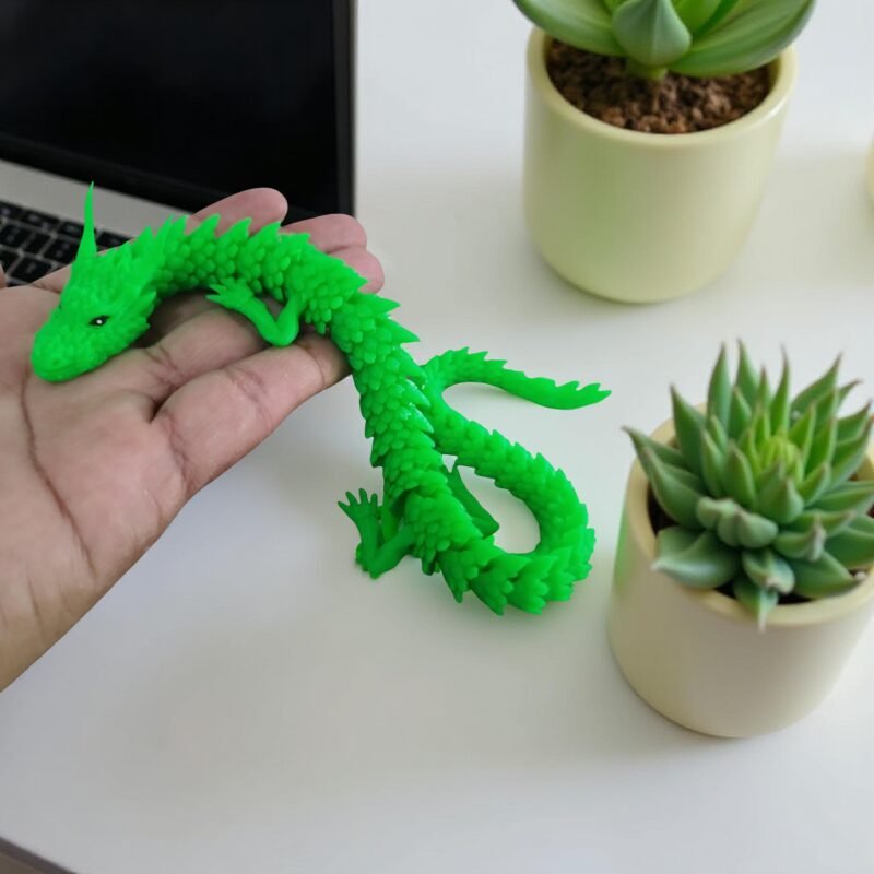 Dragão verde impresso em 3D sendo segurado na mão, com plantas suculentas ao fundo